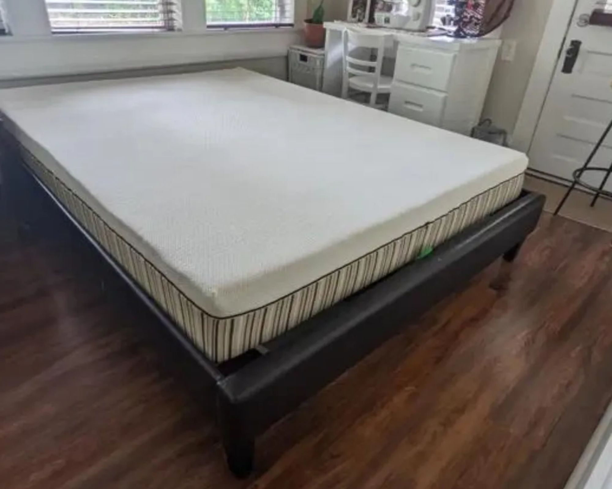 A white essentia Rem5 Mattress on a low bedframe in a white bedroom. The flooring is warm wood, and above the bed the bottom portion of two windows with slatted blinds can be seen. To the right of the bed is a white desk with a matching chair, and a white door.