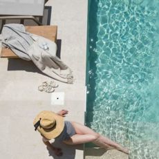 Woman wearing sunhat with legs in pool, sun lounger and towel nearby.