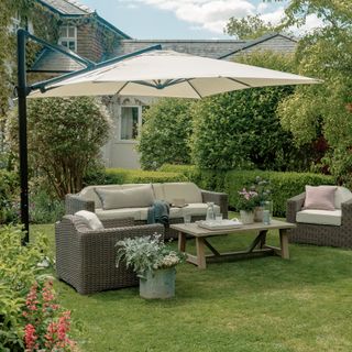 Wicker outdoor sofa and armchair set with wooden coffee table set out on a lawn below a large square cream parasol with black pole