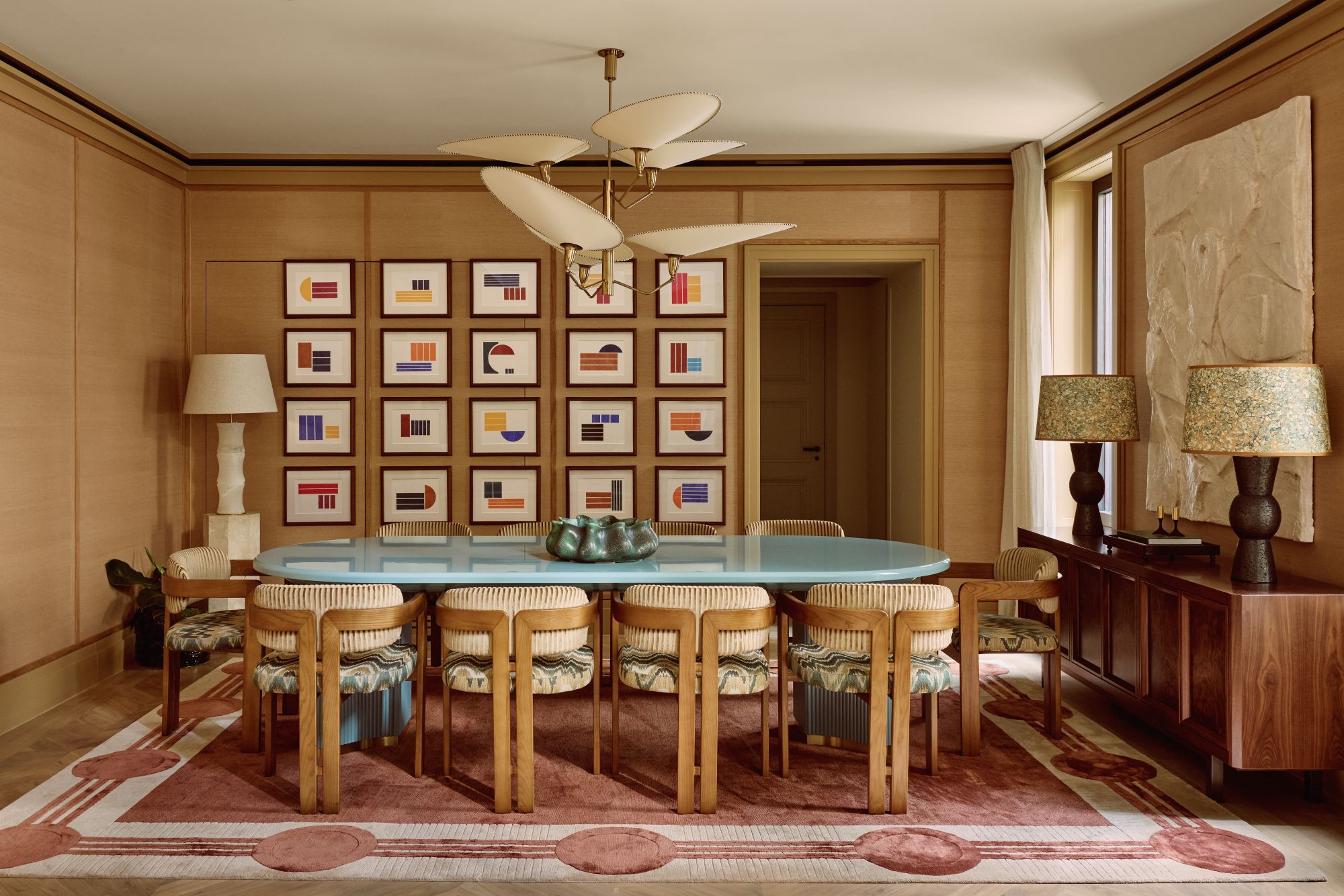 a large dining room with a baby blue lacquer table, several dining chairs flanking the sides, a credenza with two dark wood lamps, and a wall of several abstract prints framed