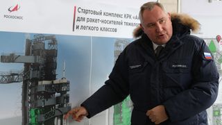 dmitry rogozin, wearing a coat, pointing to a wall poster of a rocket inside of a launch tower