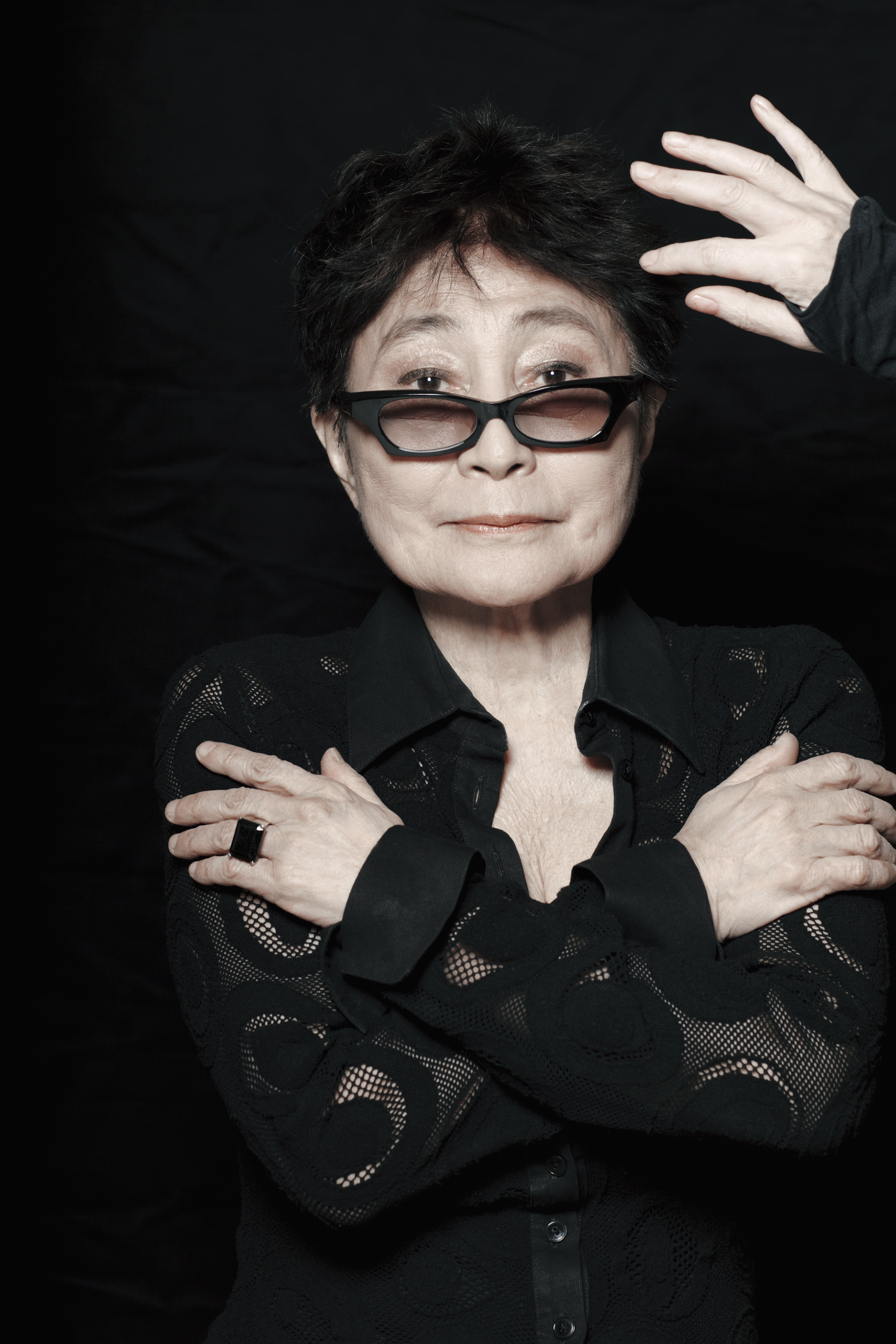 Yoko Ono faces the camera with arms crossed and one hand raised against a black background, wearing a black lace shirt and distinctive cat-eye glasses with tinted lenses.