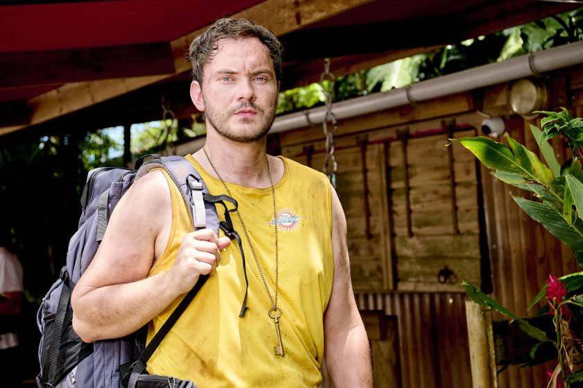 Jonny Feldon (Simon Lennon) stands wearing a yellow sleeveless top with the &quot;Island Warrior&quot; logo on it, in front of a wooden cabinet outdoors surrounded by greenery, and under a wooden canopy. He has a grey rucksack slung over his right shoulder, and his face, arms and top are all quite dirty, suggesting some sort of intense outdoor physical activity. He is also wearing a key on a chain around his neck.