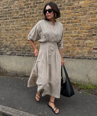 British influencer Lucy Alston walking on a London sidewalk wearing a neutral cinched-waist shirtdress, a black shoulder bag, and black strappy flat sandals.