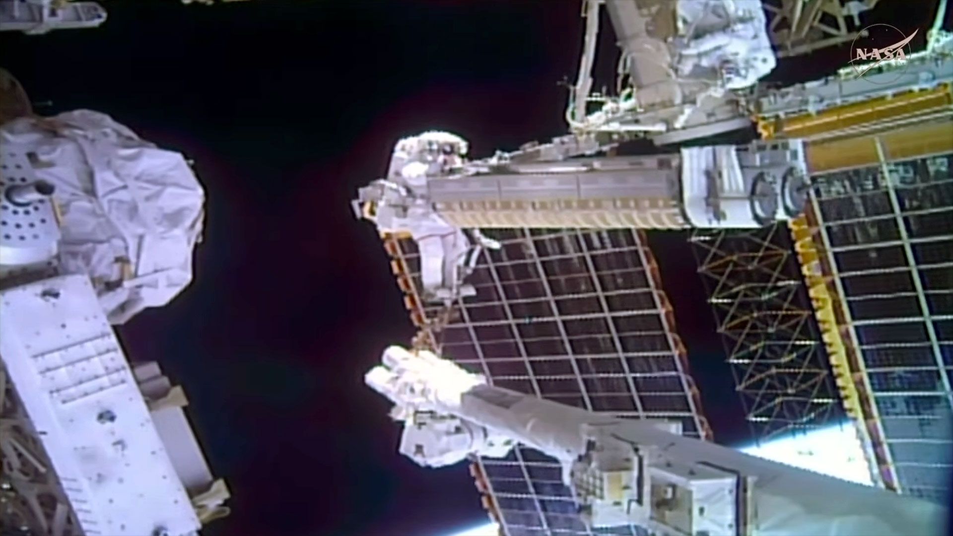 a spacesuited astronaut is seen holding a large piece of cargo while riding at the end of a robotic arm outside of a space station