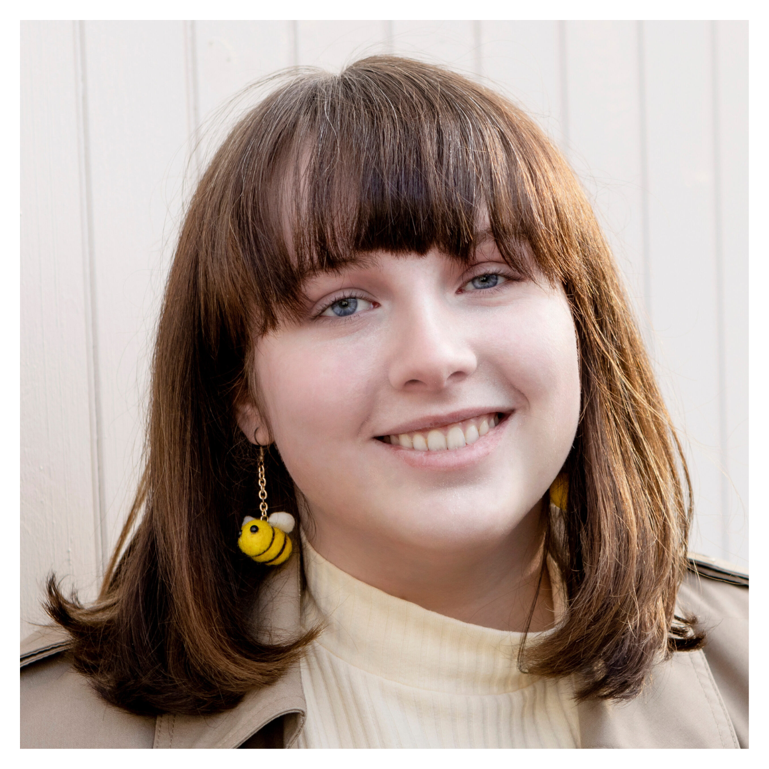 A headshot of a girl with a brown lob and bangs in a cream high-neck sweater and a coat with a dangling bumblebee earring