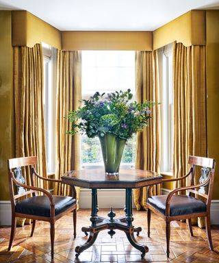 Classic living area with dark wooden table and chairs, mustard painted walls and matching curtains with pelmet on large window area, large vase with flowers on table