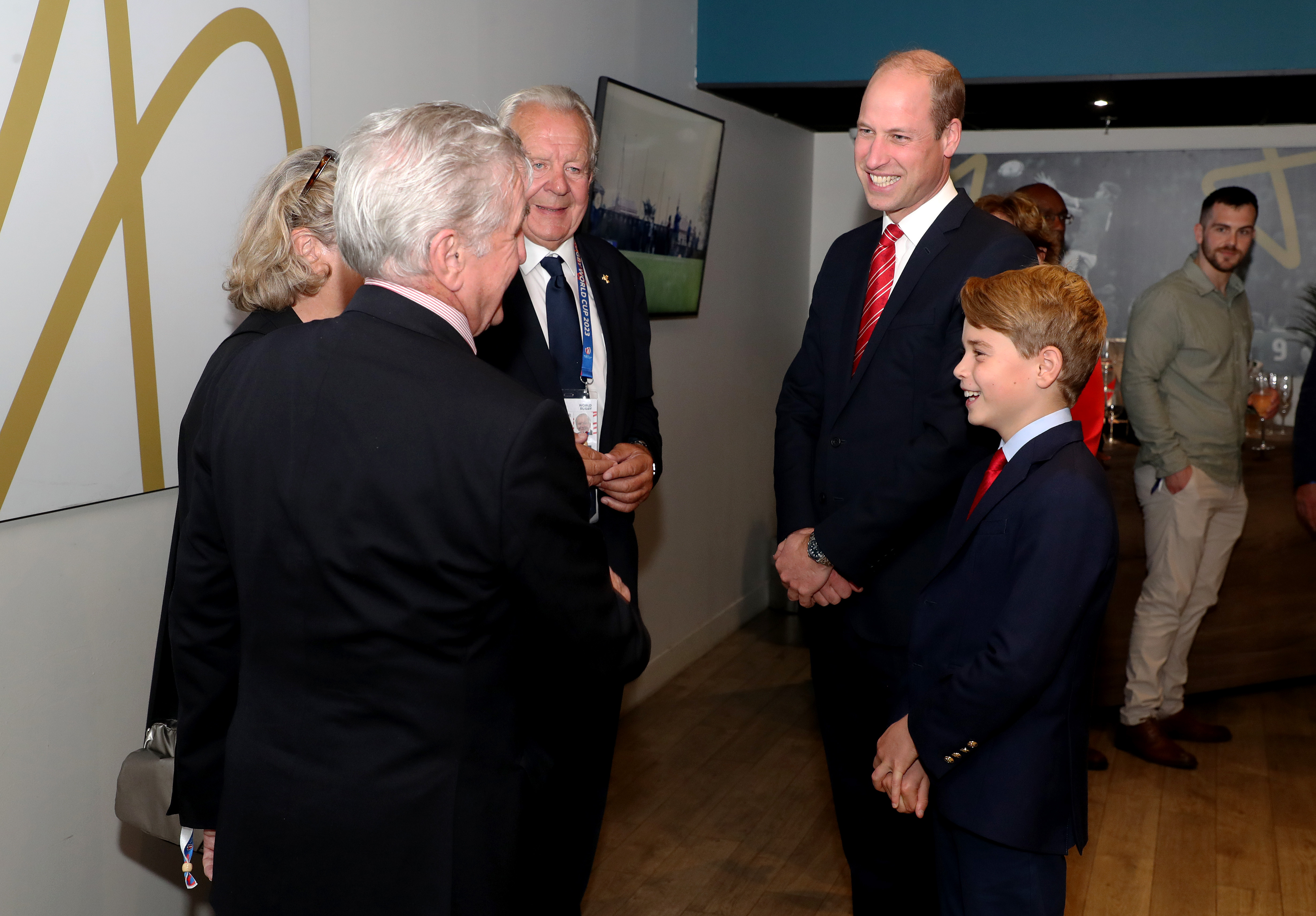 Prince George and Prince William wearing suits and smiling talking to a group of people