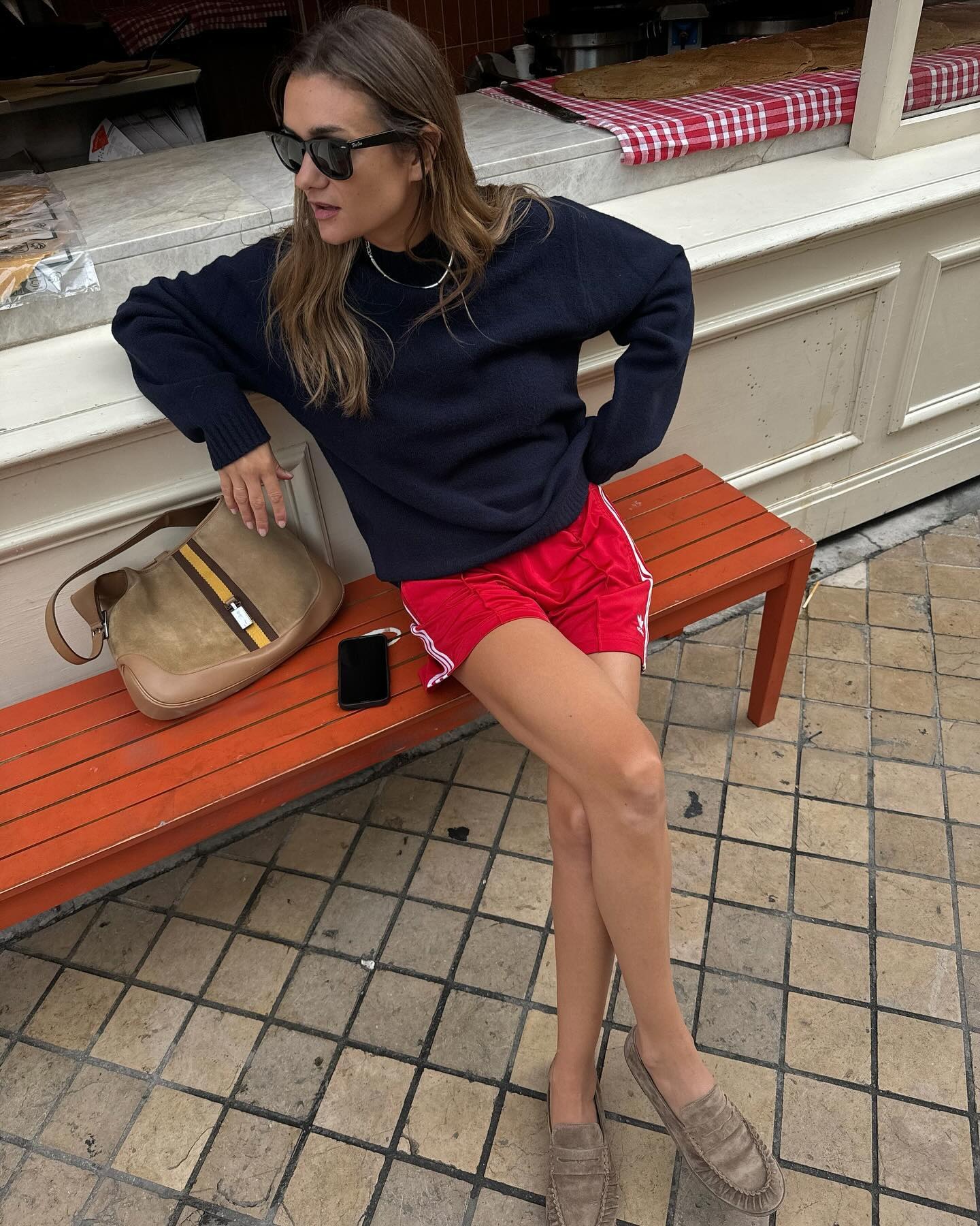 French fashion influencer Anne-Laure Mais sites on a red bench outside a Paris cafe wearing black sunglasses, a navy blue sweater, red Adidas track shorts, and tan suede loafers