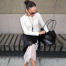 Style influencer Ingrid Fagerli Edvinsen poses in Olso, Norway wearing a white sweater, long black pendant necklace, a black woven bag, black slip skirt with contract white lace hem