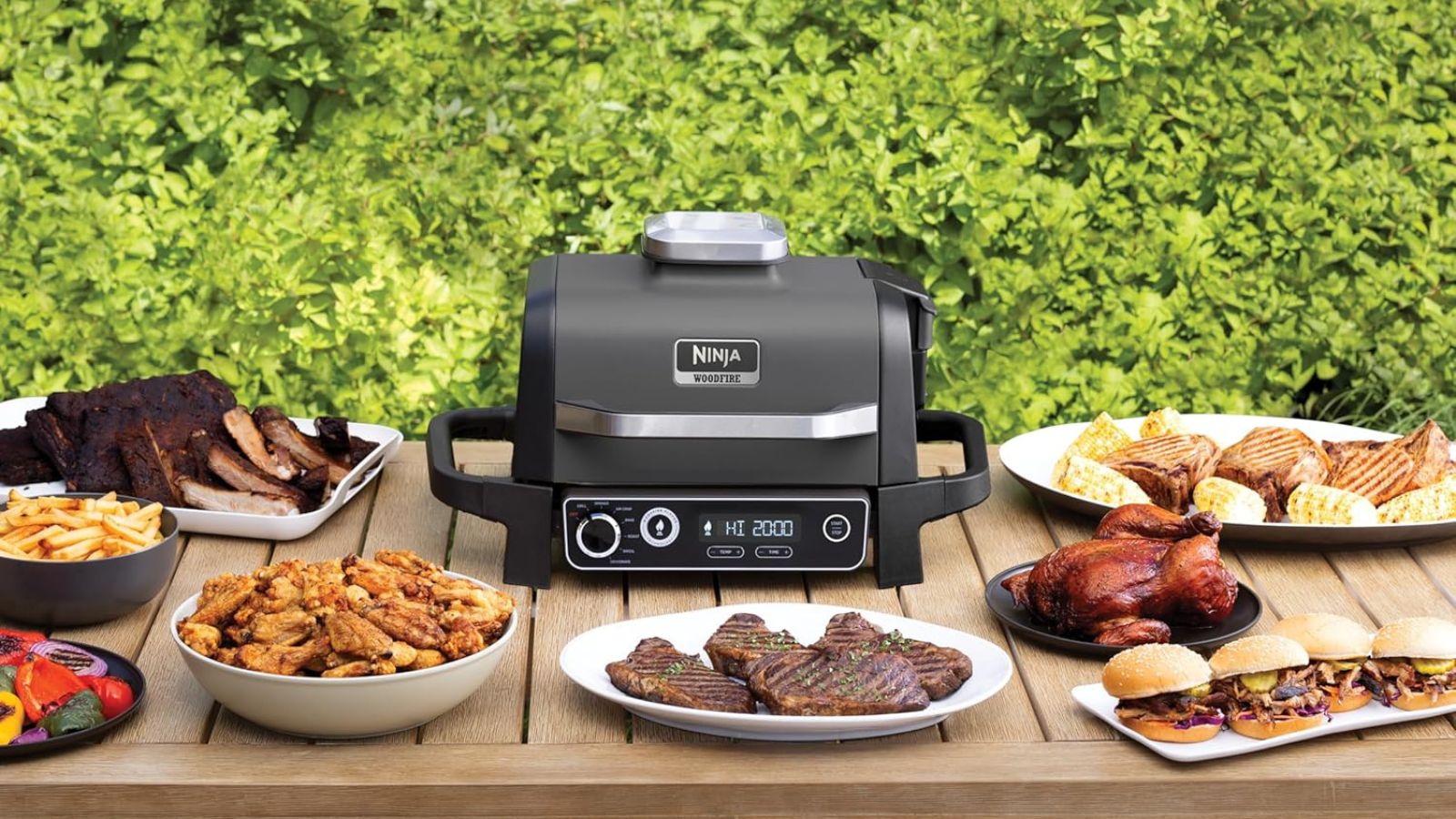 Ninja Woodfire outdoor grill surrounded by plates of meat and grilled food on wooden table, in front of green bush.