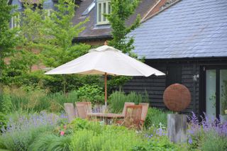 A backyard dining area with a wooden table and chairs and a cream parasol surrounded by green and purple plants