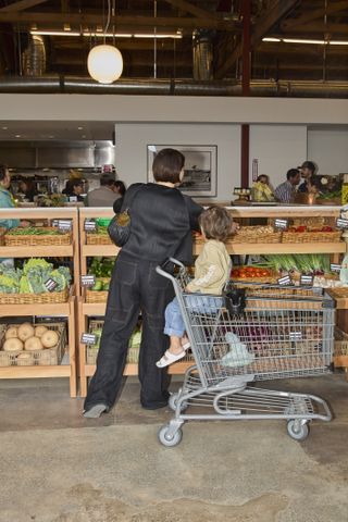 A woman dressed in a black top and trousers and a child wearing white sandals, jeans, and a cream top browse for groceries facing the camera with their back at a supermarket with industrial decor.