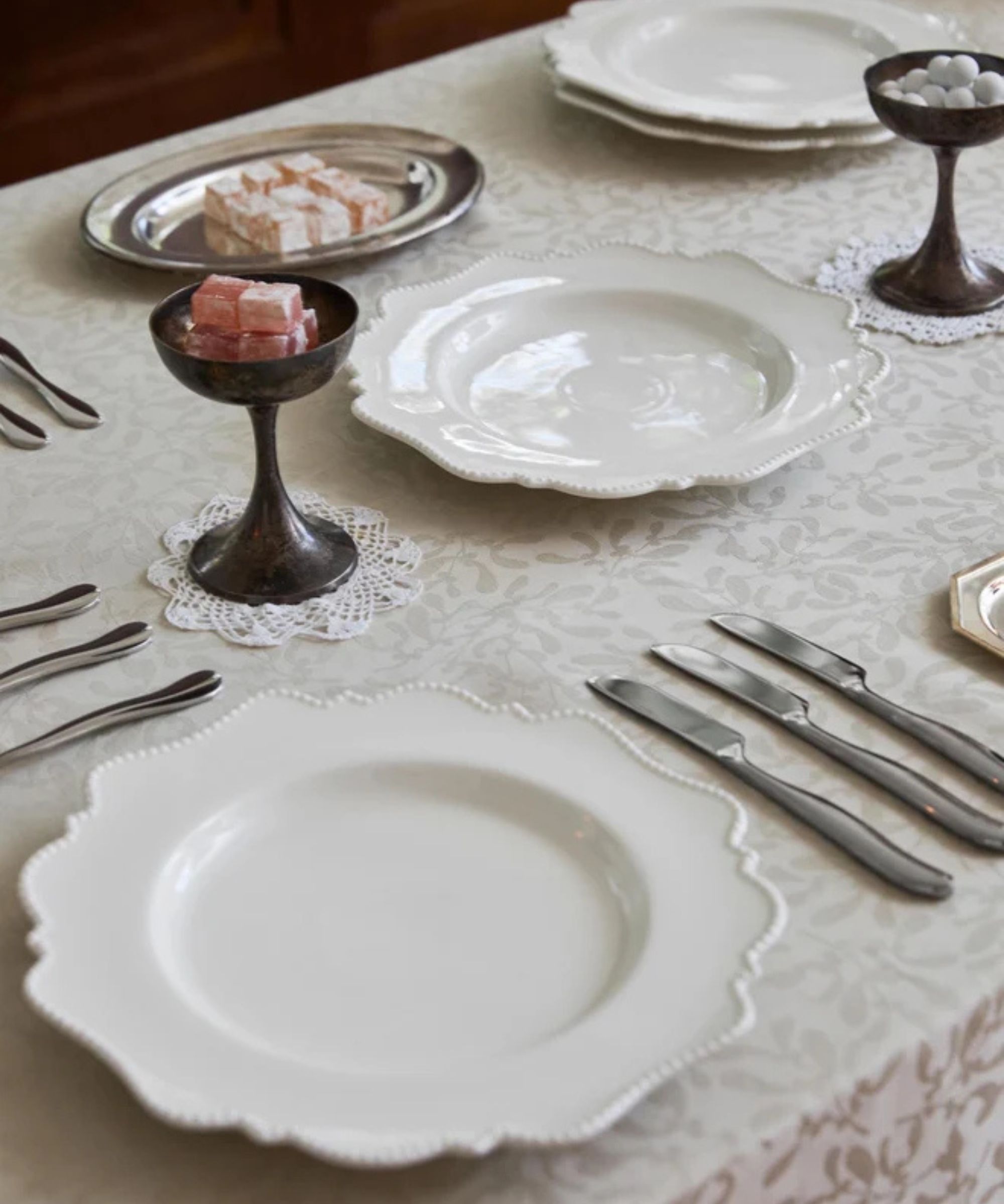 table set with white patterned tablecloth, silver cutlery, scalloped white dinner plates, doilies, silver plates