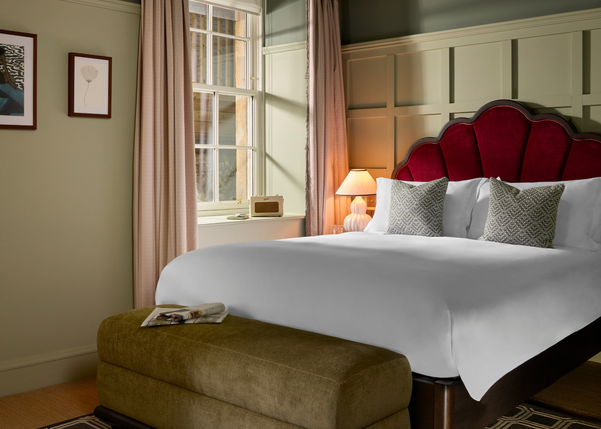 A pale green bedroom with beige window treatments, a bed with a red scalloped headboard, a green storage bench, and patterned pillows by a ribbed white table lamp