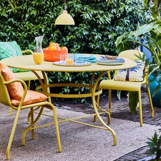 yellow painted table and chairs in garden, matching pendant light, bowl of oranges, rug, bright cushions, cutlery, plates, jug of orange