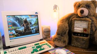A retro PC encased in a jumbo Teddy Bear on a desk next to a CRT monitor outputting artwork of a bear in the wild and an IBM model M keyboard with some green highlight keys.