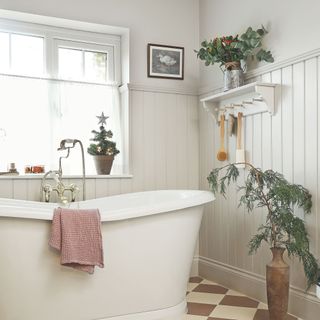 White bathroom with white panelling on the wall, a freestanding bath and cafe curtains over the bathroom window