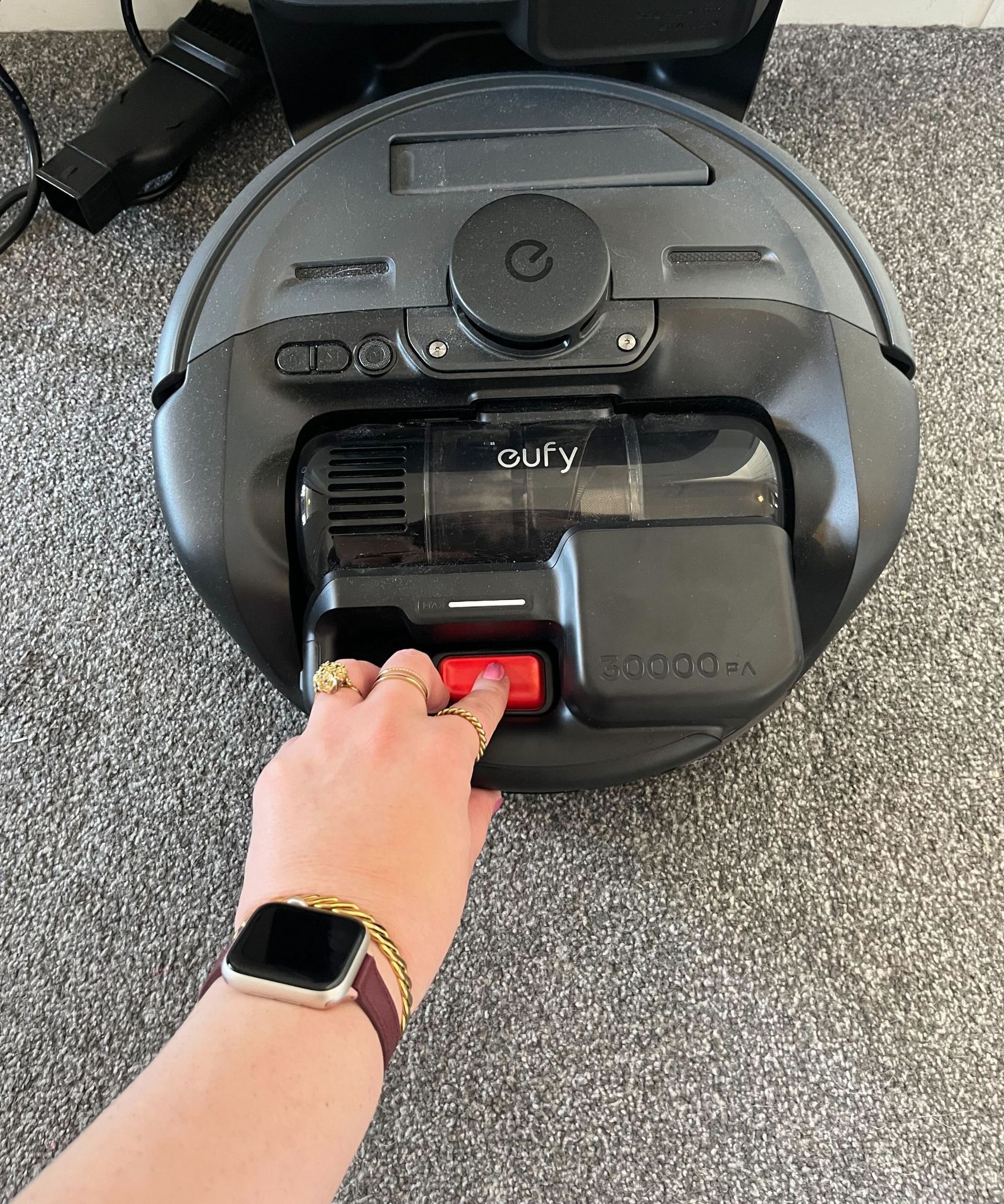 A black round robot vacuum is on a carpeted gray floor. There is a hand pushing the red release button location on the top