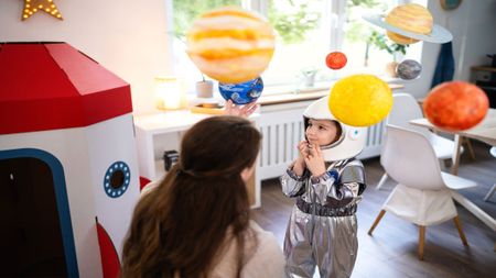 A Kid wearing an astronaut costume is looking at model planets with a parent.