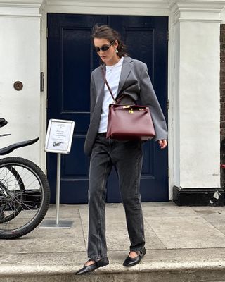 Influencer fesyen melangkah keluar dari trotoar mengenakan pakaian penuh gaya dengan kacamata hitam oval, blazer abu-abu, kaos putih, tas Hermès merah anggur, jeans hitam pudar, dan sepatu flat Mary Jane hitam.