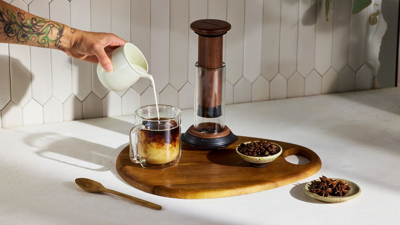 Person pouring milk into coffee on wooden board beside Walnut AeroPress coffee maker