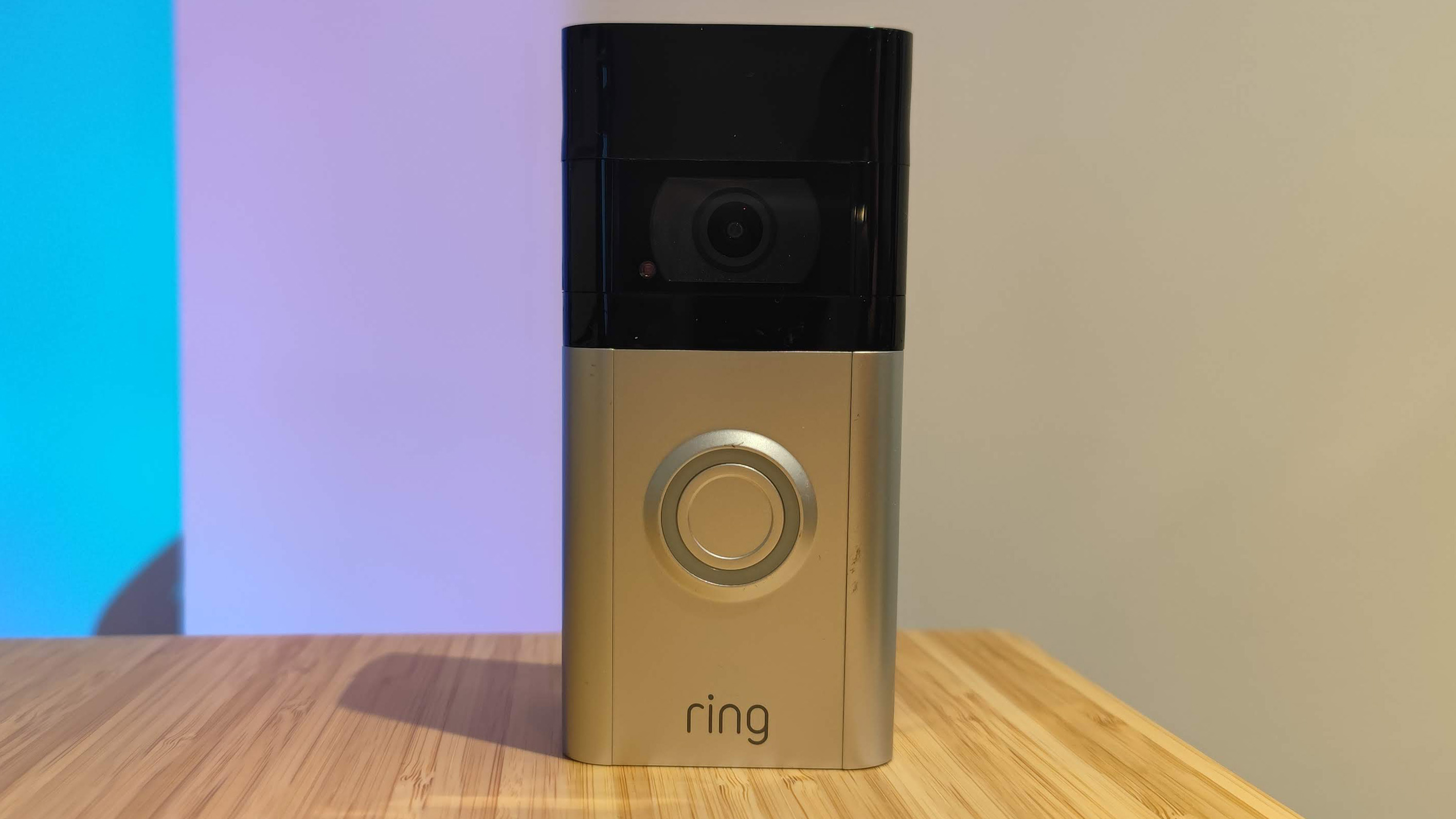 En svart och silvrig Ring Video Doorbell 4 står på ett träfärgat bord mot en vit vägg som lyses upp av blått och lila ljus.