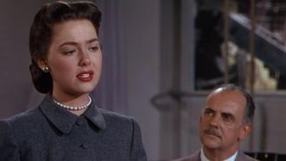 A man, sitting down, looks quizzingly up at a woman, standing up, wearing white pearls and red lipstick