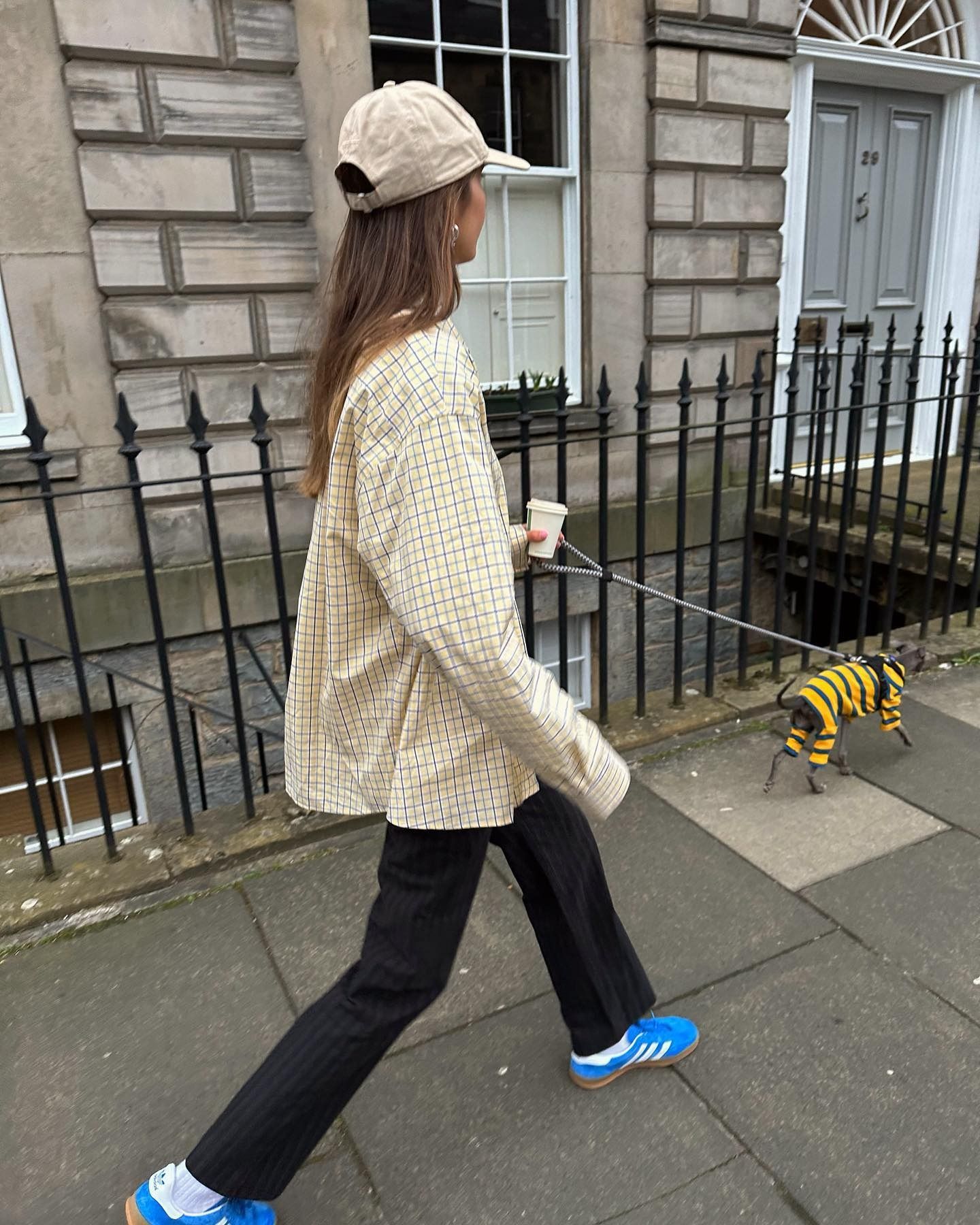 British style influencer Poppy Almond walks her Italian Greyhound dog wearing a baseball cap, checkered button-down, pinstripe flared pants, and bright blue Adidas Gazelle sneakers