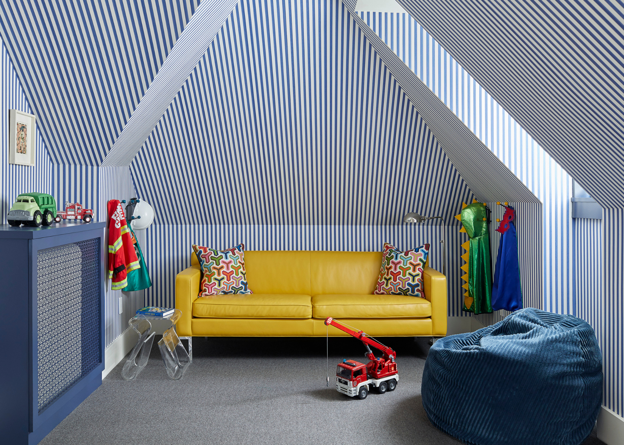 A playroom space with sloped ceilings and walls covered in blue and white striped wallpaper with a yellow sofa and a teal beanbag in front as well as a blue radiator cover with toys on top and on the floor