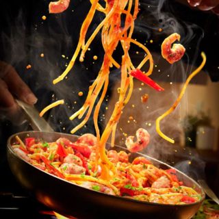 A lifestyle image of a pan full of noodles and prawns being tossed in the air during cooking on a black background