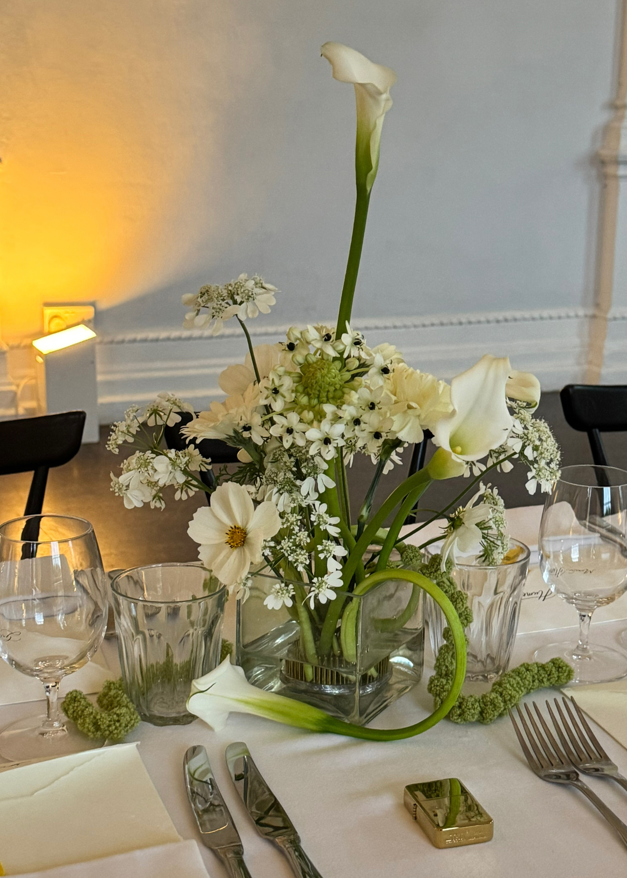 A table set with white linens, a gold lighter, silver cutlery, and a white floral centrepiece with calla lilies