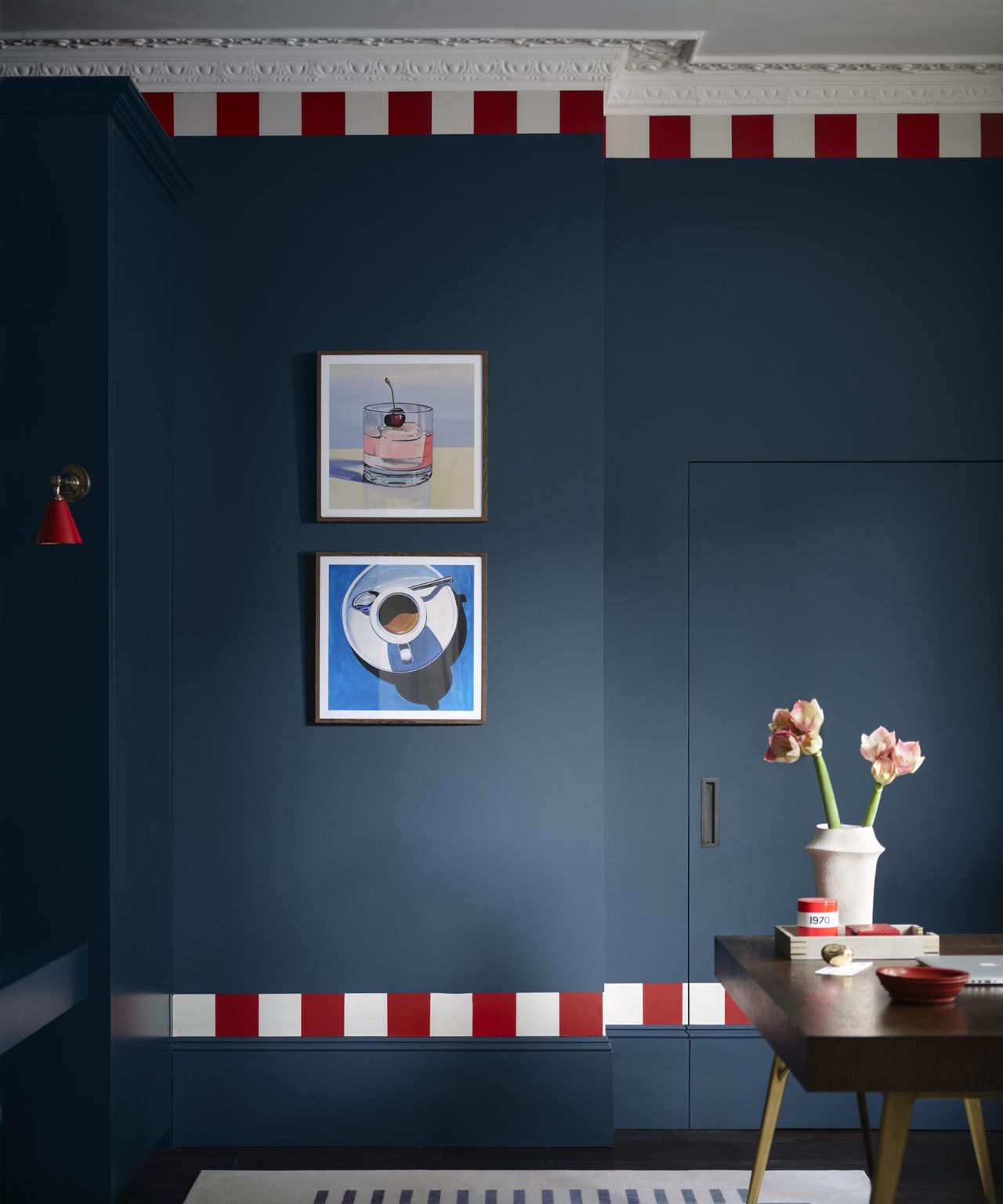 A dramatic navy blue room with red and white striped detailing on the skirting and cornice. Modern artwork hangs above a dark wooden dining table, creating a striking, contemporary contrast.