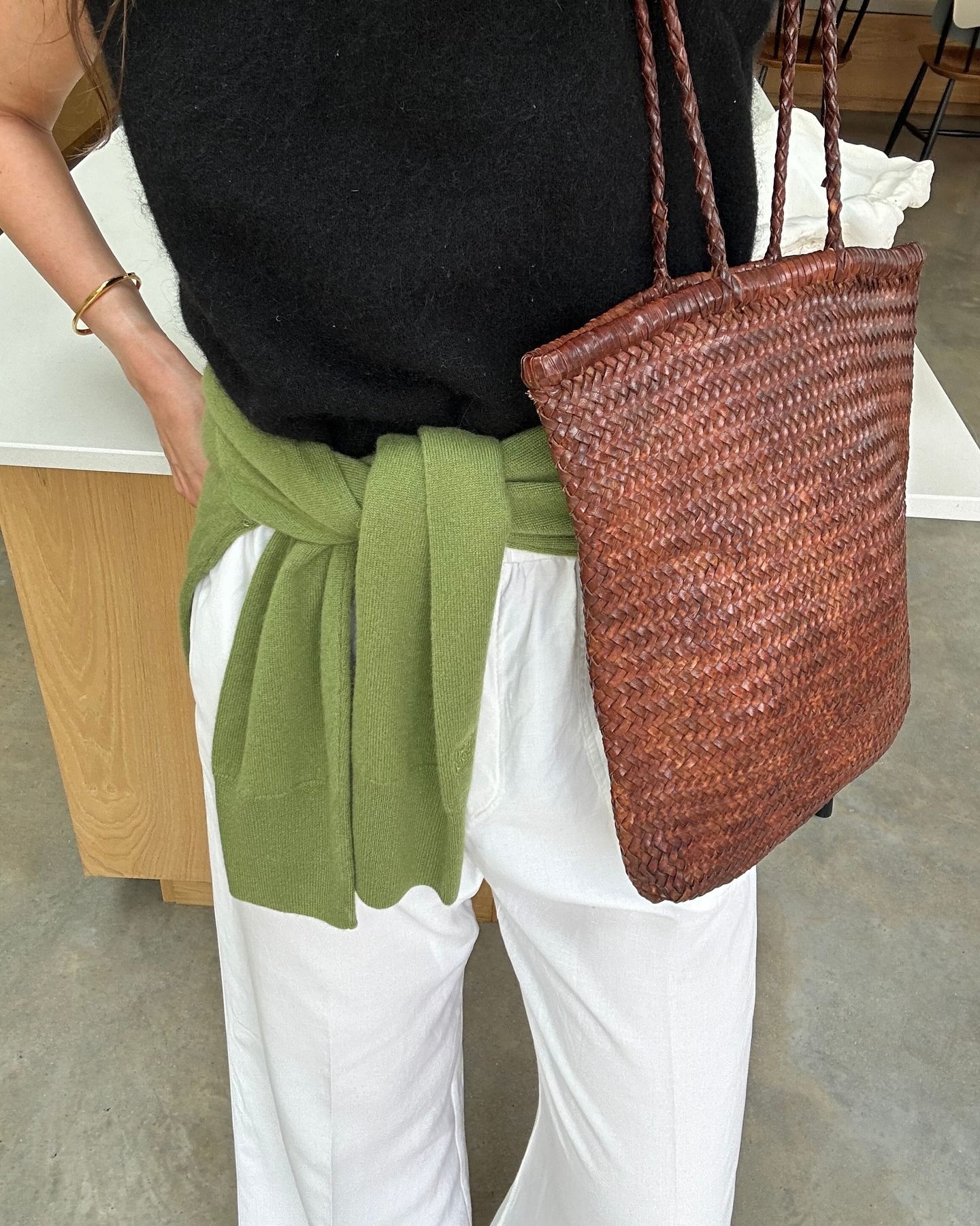 British style influencer Anna Howard poses with a woven brown St. Agni tote bag wearing a knit sleeveless top, green sweater tied around the waist, and white linen pants.