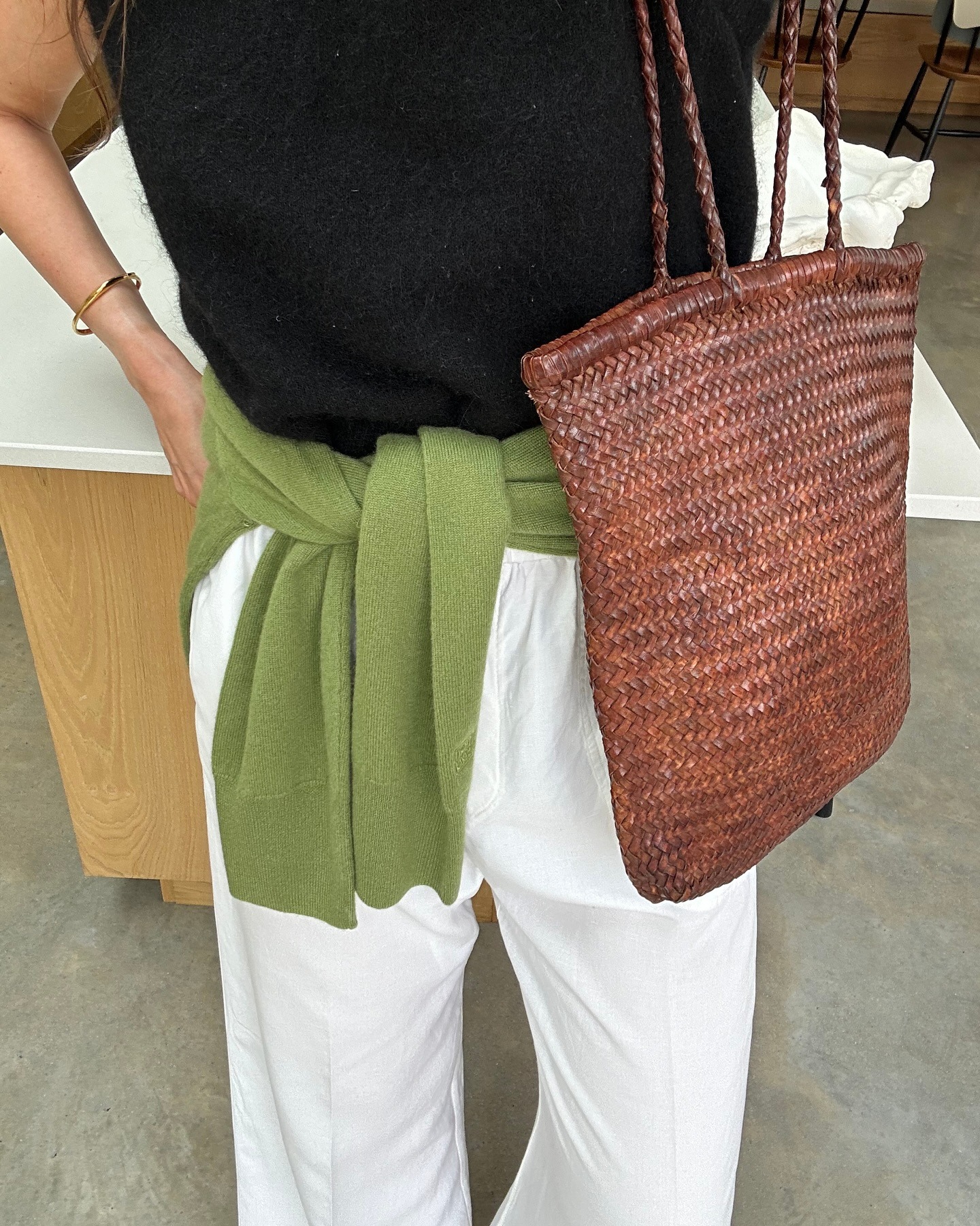British style influencer Anna Howard poses with a woven brown St. Agni tote bag wearing a knit sleeveless top, green sweater tied around the waist, and white linen pants.