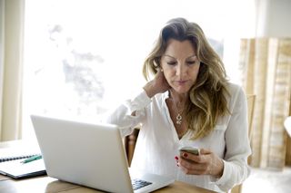 Menopause and job loss: Mature woman using laptop whilst reading smartphone text at home