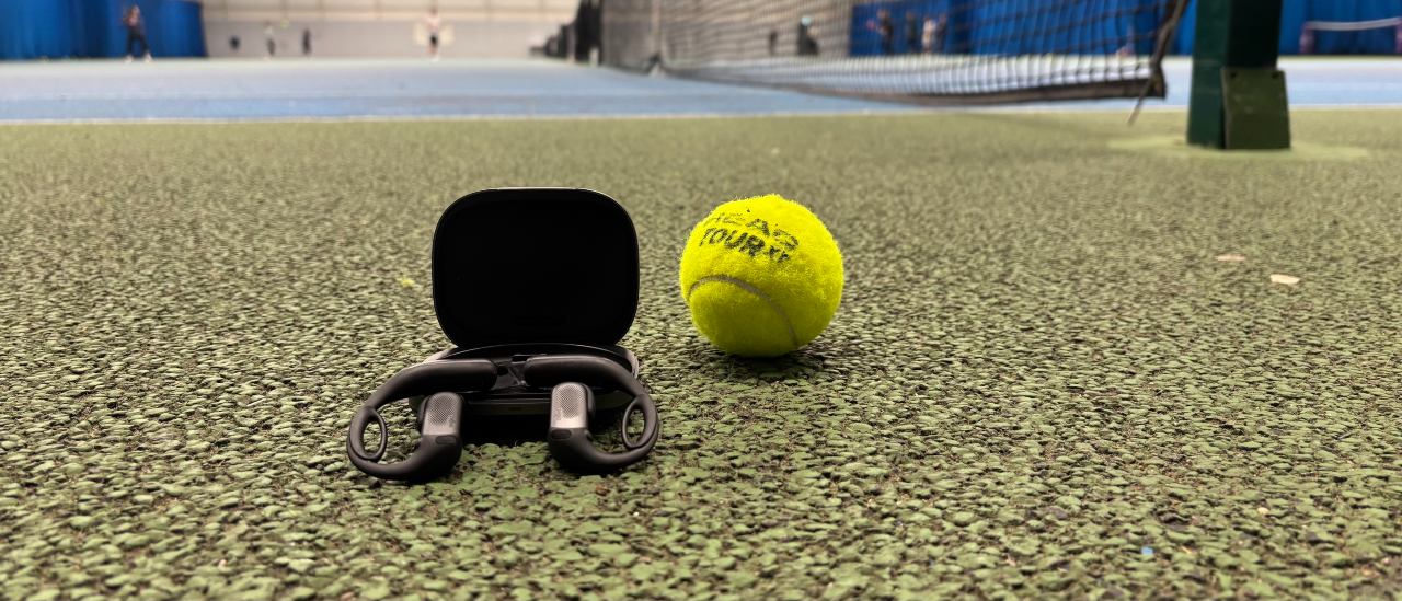 Shokz OpenFit Pro open earbuds, balancing on their carry case, next to a tennis ball at an indoor tennis center.
