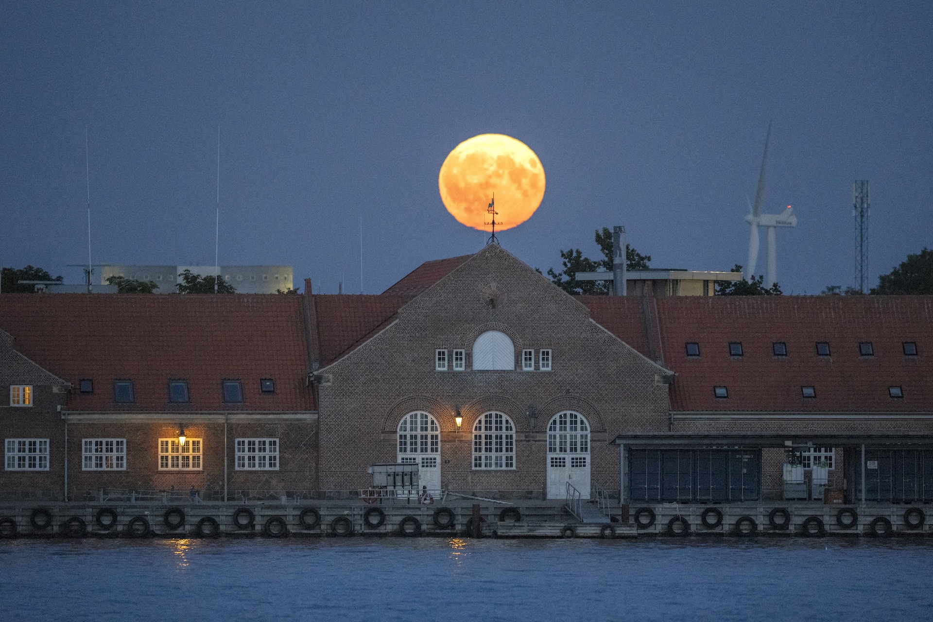 A yellow moon rises behind a building on the water