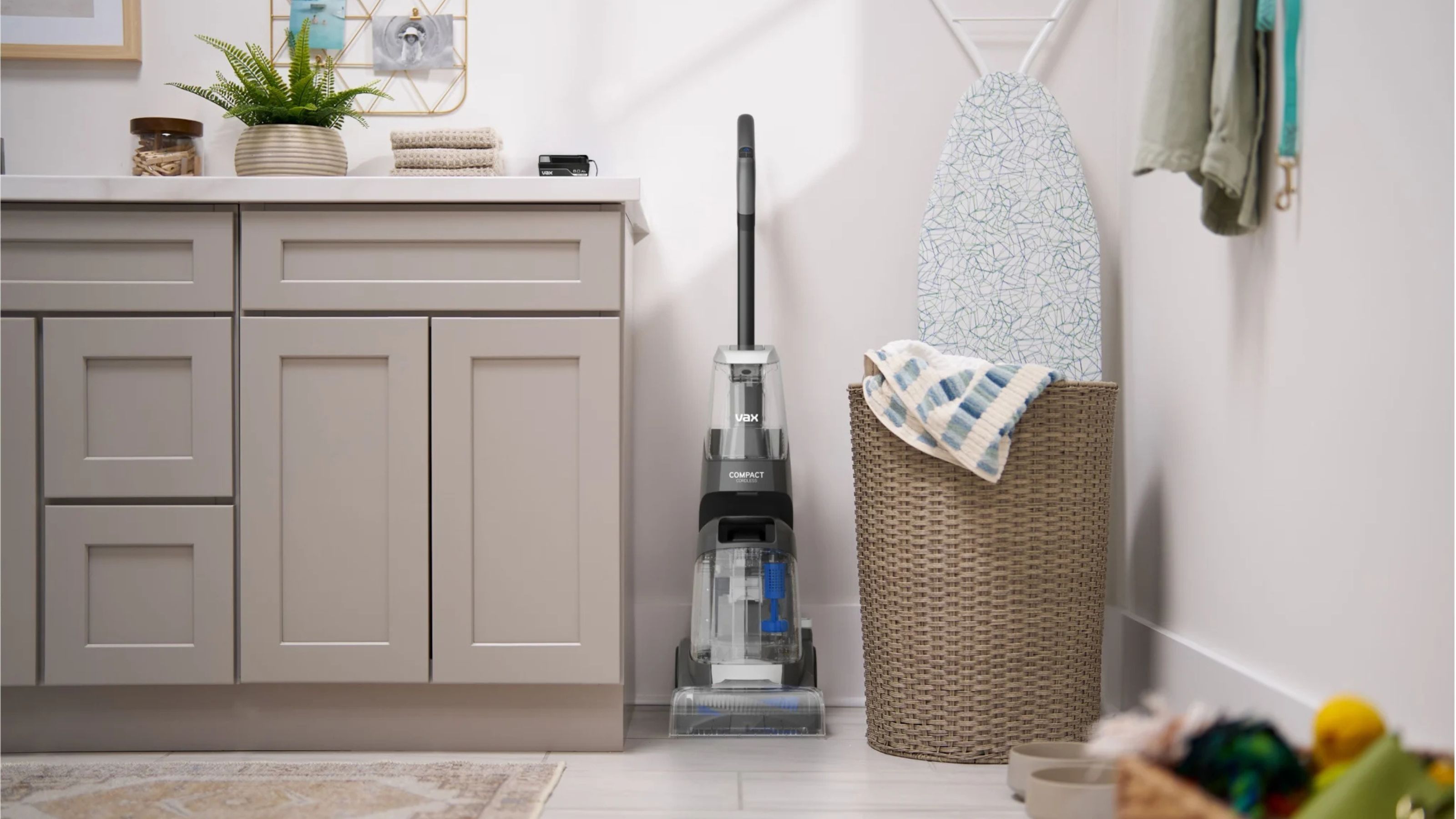VAX ONEPWR Compact Cordless Carpet Cleaner in a utility room next to an ironing board, laundry basket, and cabinets