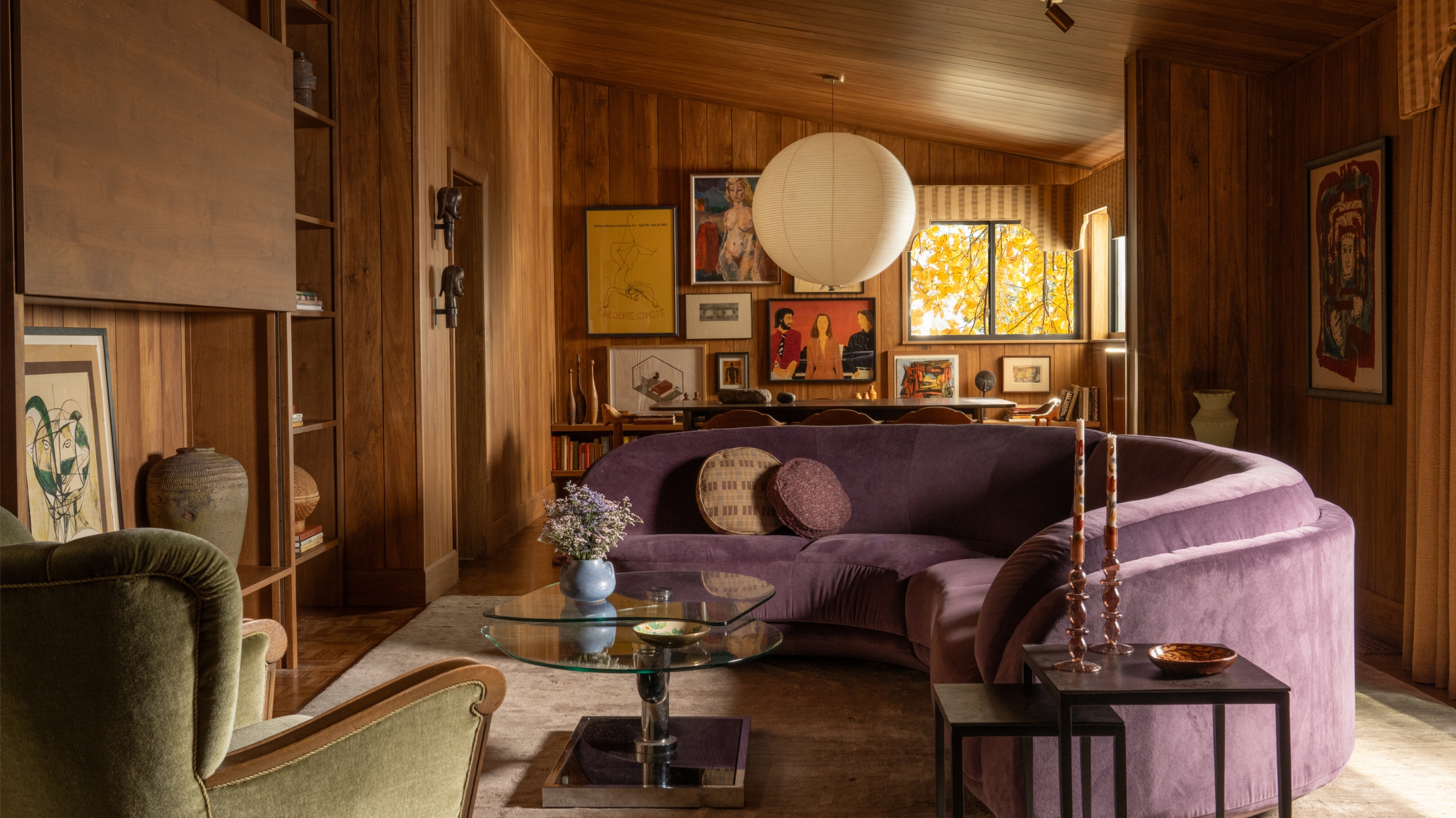 A mid-century living room with wood paneled walls and sloped ceiling, a curved purple sofa, and round paper pendant
