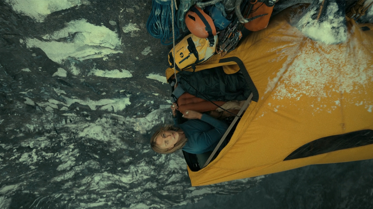 Charlize Theron sticking her head out of a tent attached to the side of a cliff in Apex