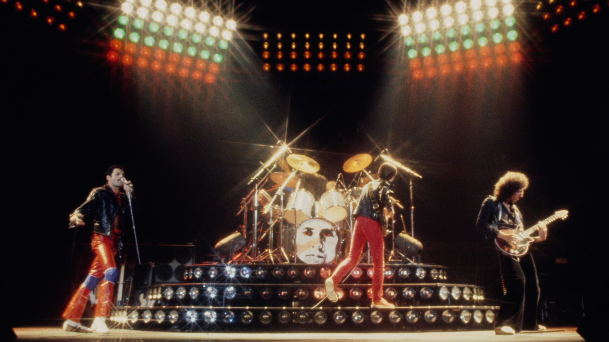 British singer, songwriter and musician Freddie Mercury, wearing a black leather jacket and red leather trousers, British bass guitarist and songwriter John Deacon, who wears a black jacket and red trousers, and British guitarist and songwriter Brian May, in a black shirt and black trousers, as British rock band Queen perform live in concert, circa 1980