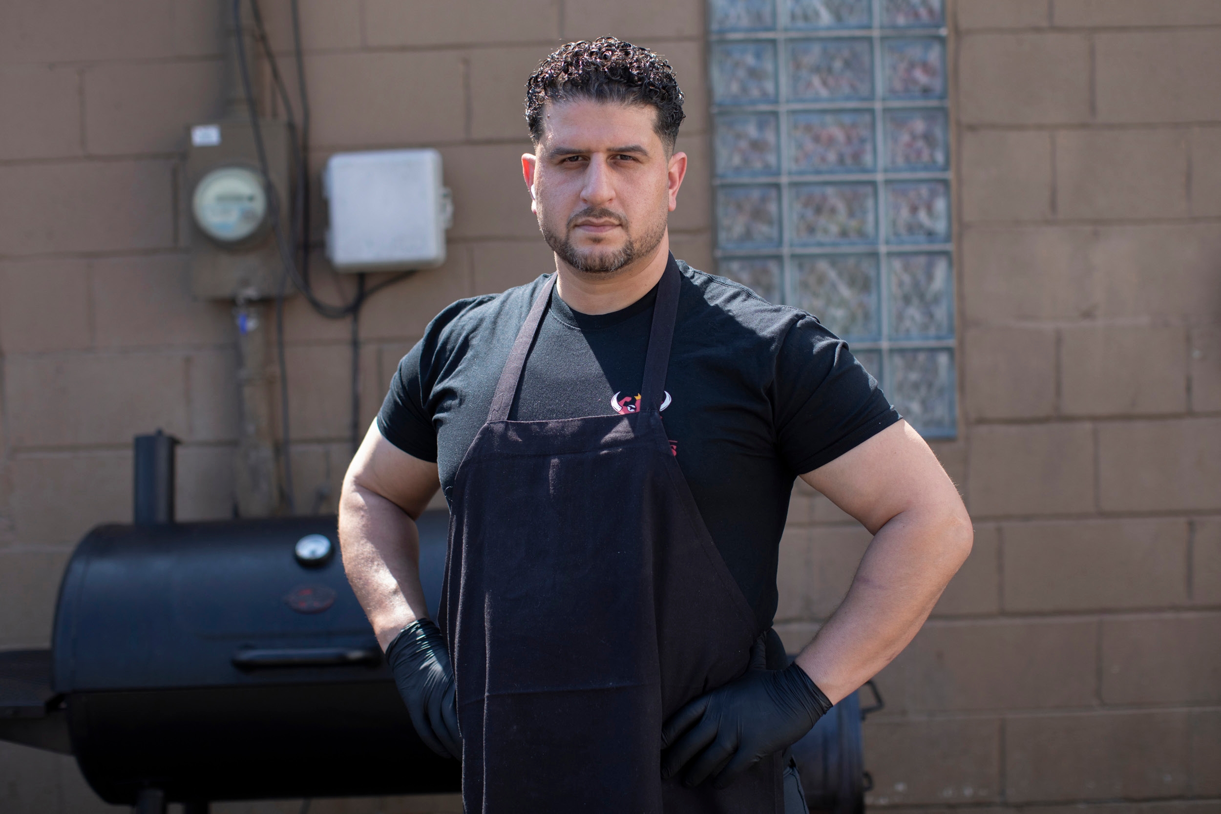 A stocky man in a black t-shirt and dark apron stands with his hands on his hips in front of a large smoker grill, looking directly at the camera against a plain brick wall.