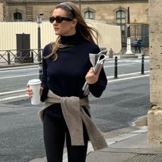 Anne-Laure Mais wearing a black turtleneck sweater, black leggings, white socks, loafers, and a tan sweater tied around her waist while walking with a coffee in Paris.