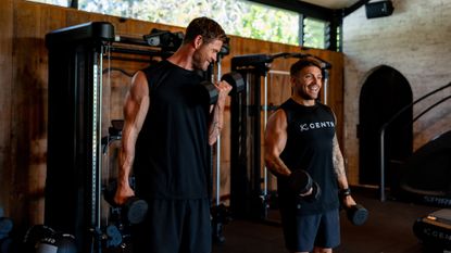 Two muscular men holding dumbbells with gym equipment behind them