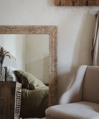 A bedroom with greige walls, a gray armchair, and wooden engraved mirror reflecting a sage green bed and dark wooden nightstand with dried flowers on