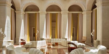 An historical palace blessed by sunshine boasts a vaulted lounge sculpted in marble, now brought to life by modular, contemporary and curvilinear sofa designs, as well as wooden armchairs and standing lamps.