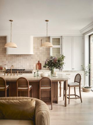 Image of a neutral and white kitchen with a large kitchen island that is wood and marble with four wooden bar seats in view. There is a pink-beige tile backsplash and two ruffled pendant lights hanging from the ceiling