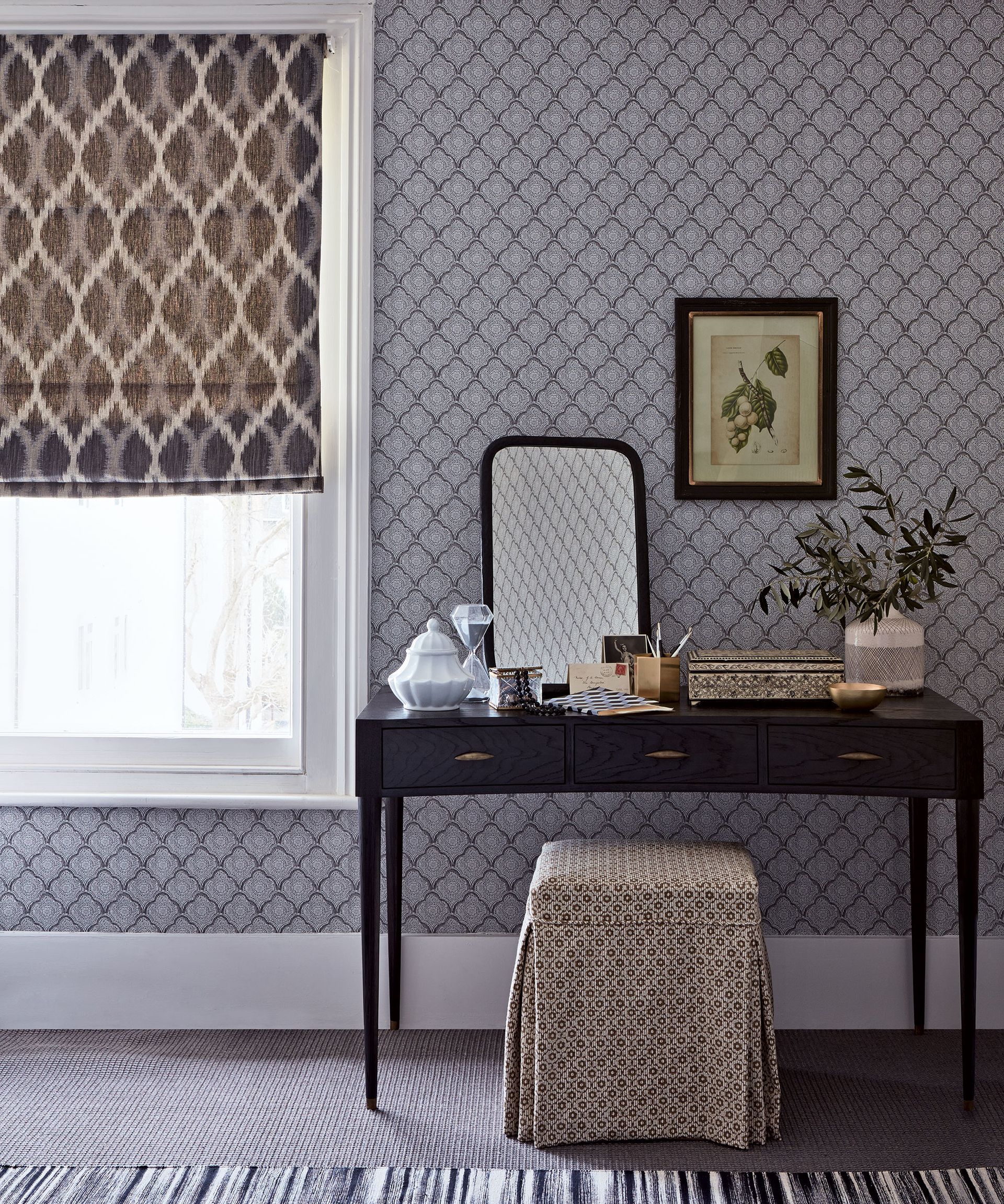 A wooden dressing table with an upholstered stool in front of patterned wallpaper