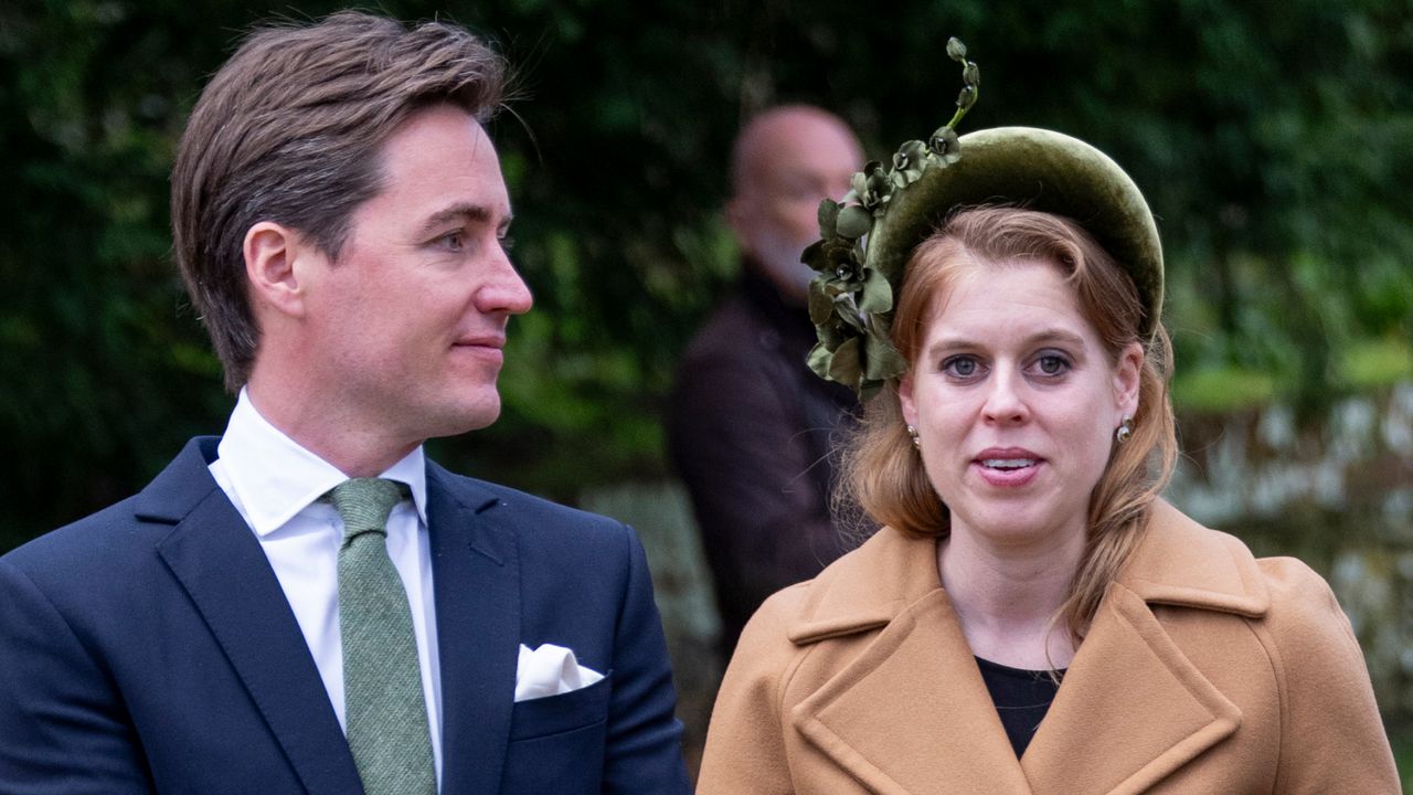 Princess Beatrice wearing a camel coat and green headband standing next to Edoardo Mapelli Mozzi wearing a navy suit and green tea outside with trees behind 